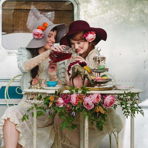 Two girls in hats enjoy a tea party for two set at a small white table with flowers.