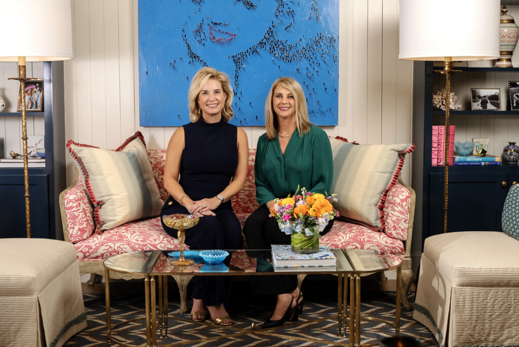 Two women are sitting on a red-patterned sofa adorned with cream pillows in a well-decorated living room. A blue painting of a woman's face is displayed on the wall behind them. There is a glass coffee table in front of them with a flower bouquet and decorative items.