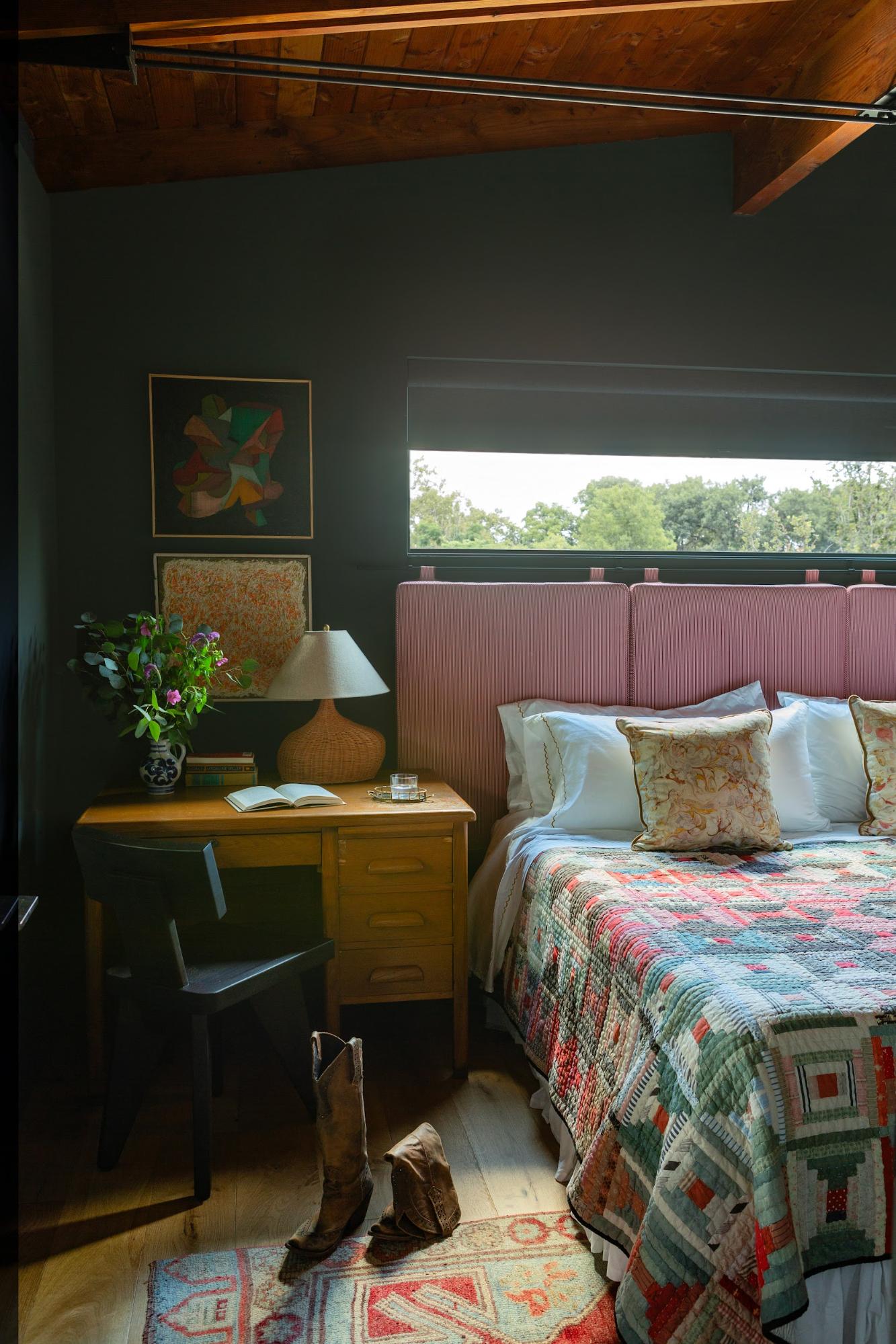 A bedroom with wooden vaulted ceilings, dark green walls, the bed made with a patterned quilt and white pillows, red pinstriped cushion headboards and a wooden side table decorated with an abstract lamp.