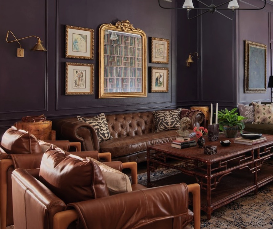 A living room with dark purple walls, a brown leather tufted sofa, two leather armchairs, a wooden coffee table, framed art, and a large mirror on the wall.