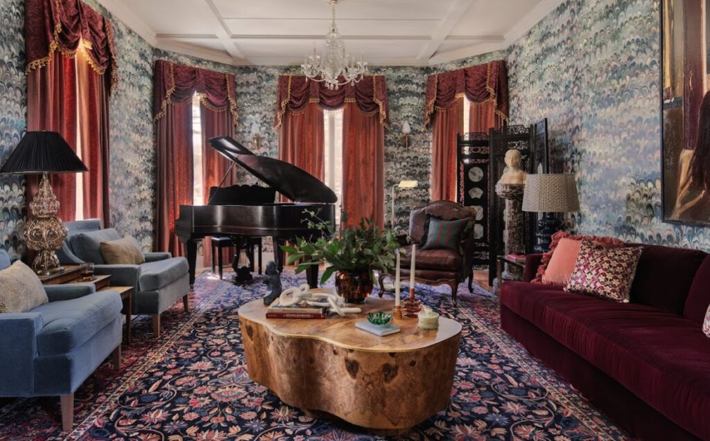 A lavish living room with ornate wallpaper, a grand piano, plush seating, a wooden coffee table, and eclectic decor including a lamp, statue, and assorted cushions.