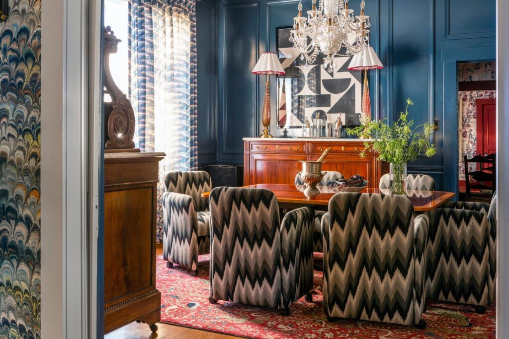 A formal dining room with a wood table, patterned chairs, a chandelier, and a sideboard. The walls are dark blue, and a patterned rug covers the floor. Decor includes lamps, artwork, and plants.