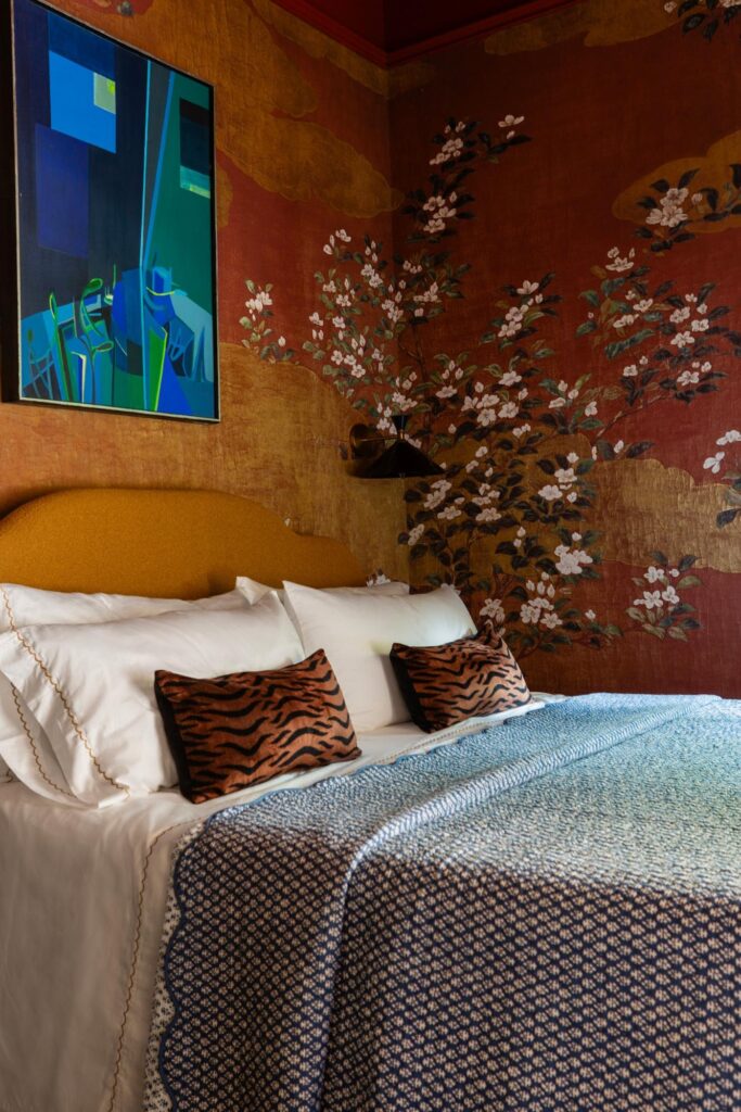 A bedroom with a bed adorned with white linens, blue pattern blanket, and tiger-striped pillows. The walls have ornate floral wallpaper, and a modern abstract painting hangs above the bed.