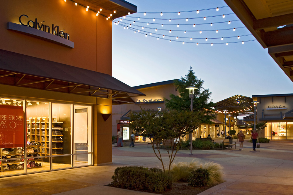 Photo of outside walking paths at the Premium Outlets in Round rock, Texas, there is shrubbery along the paths, and LED bulb light strings hung across the store fronts.