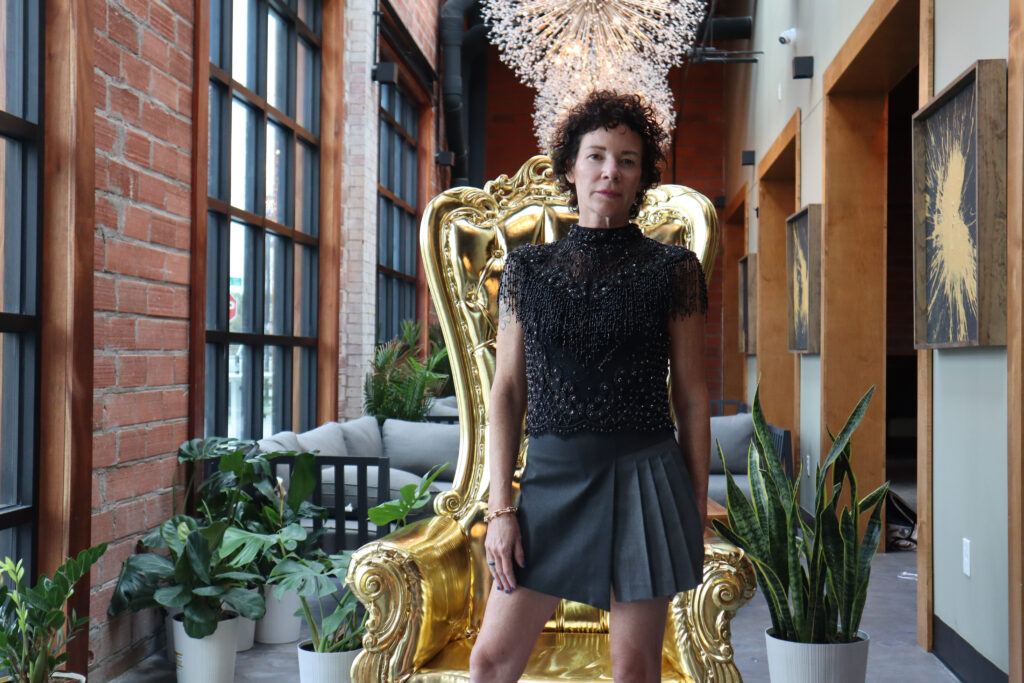 Bridget Dunlap standing in a room with elegant light fixtures and modern furniture in front of an oversized gold throne chair at the Lustre Pearl wearing a black high necked-beaded shirt and a grey pleated skirt.