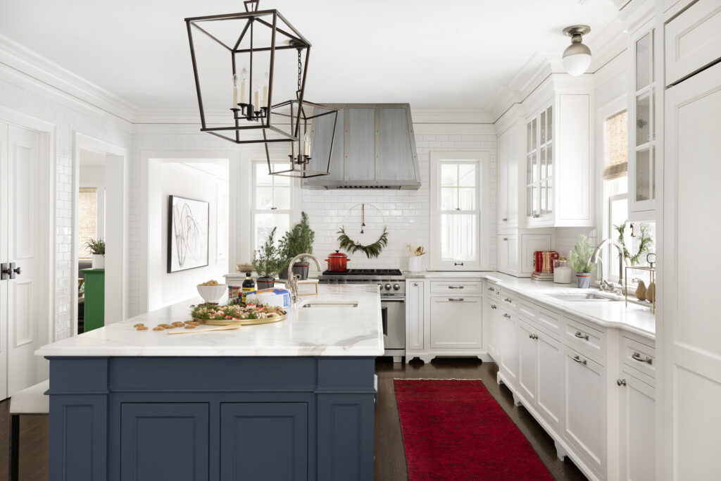 Kitchen decorated for the holidays with miniature ferns and gold serving dishes adorning the island, a crimson red runner rug and gold and leafy wreath hanging on the walls.