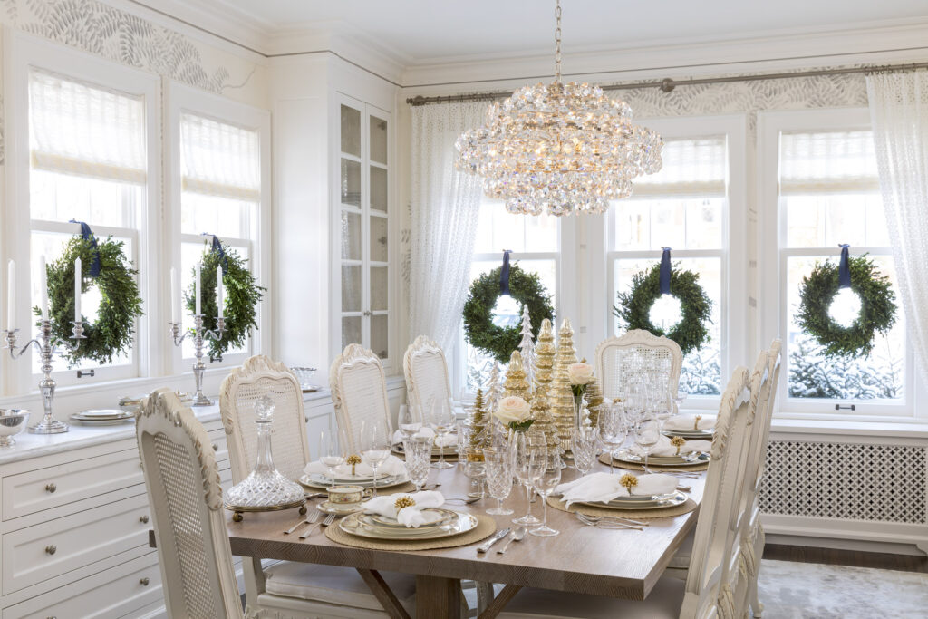 Formal dining room decorated for the holidays, five windows have wreaths hanging made out of greenery, gold miniature holiday tree sculptures, gold and white decorative plates and white roses adorn the dining table.